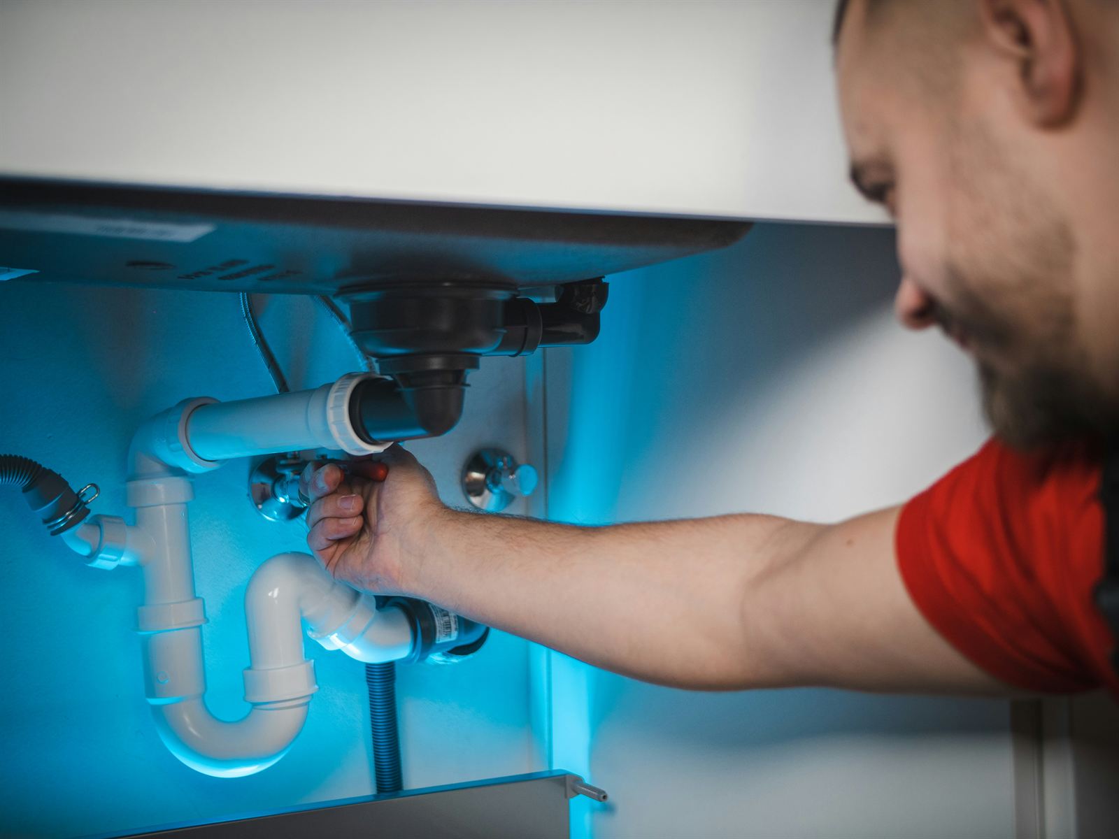 A plumber reaching under a kitchen sink to repair the pipework, lit by a work light.